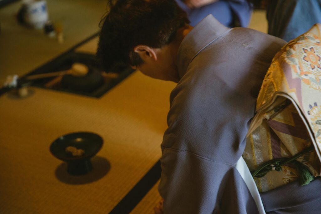 Side view of anonymous male in authentic robe bowing head while sitting on tatami with dishware for traditional tea ceremony