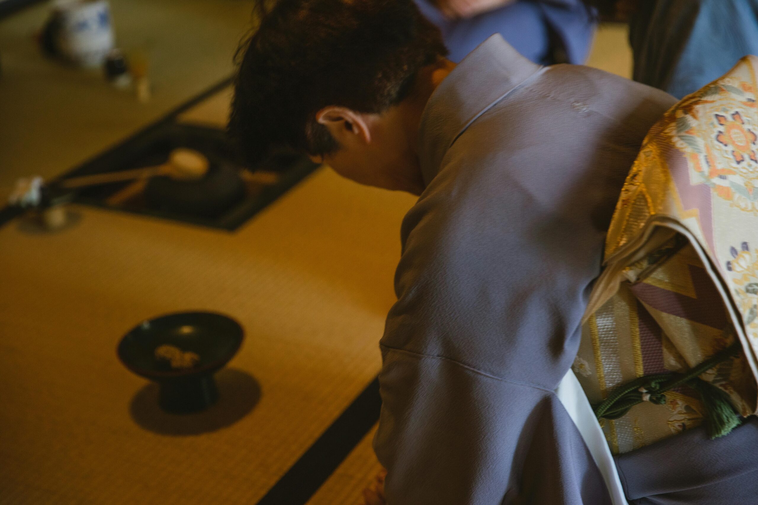 Side view of anonymous male in authentic robe bowing head while sitting on tatami with dishware for traditional tea ceremony