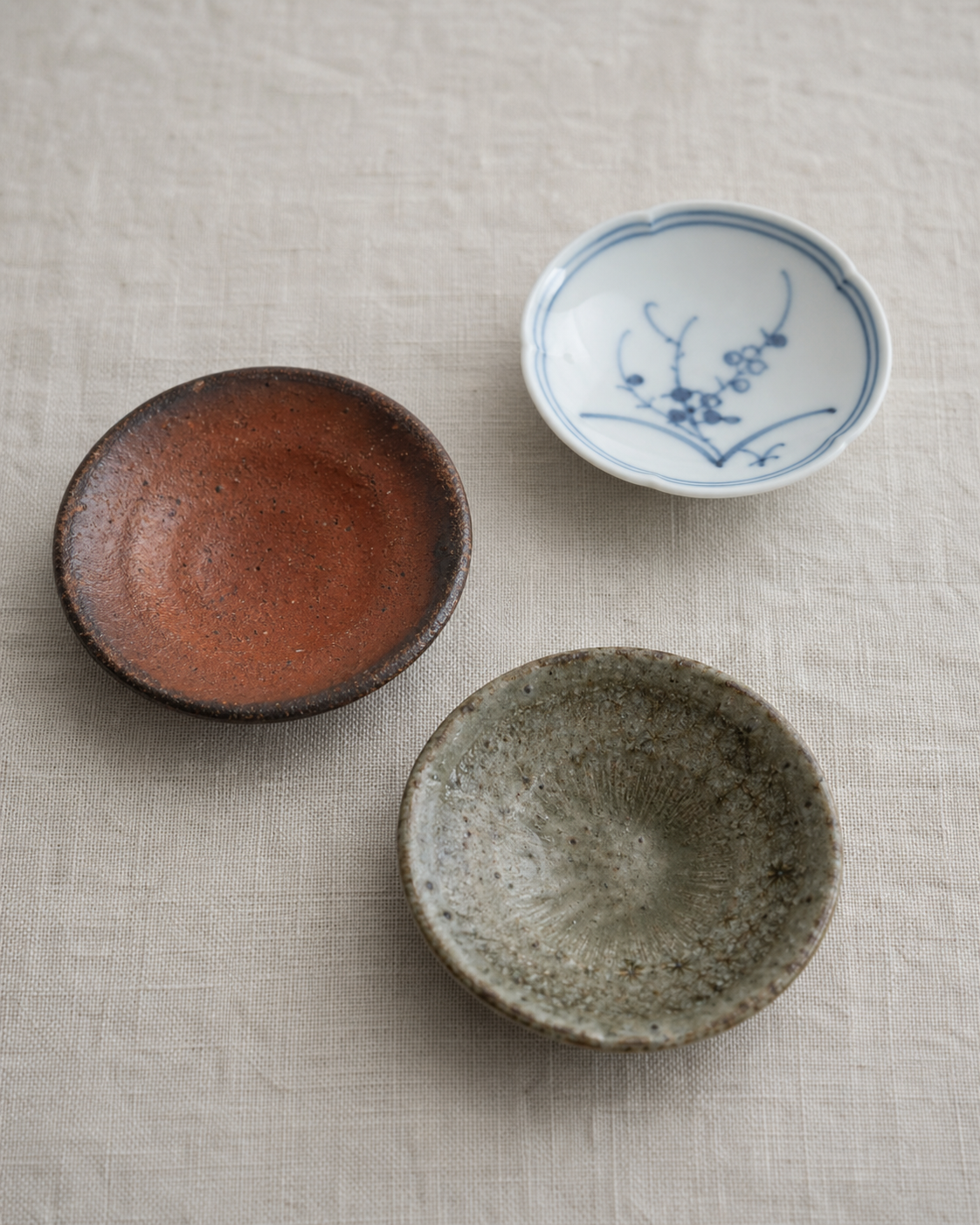 Three small Japanese ceramic mamezara plates of different kiln styles arranged on a linen cloth