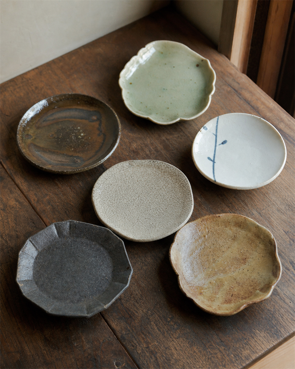 A small collection of mismatched mamezara plates on a wooden shelf, each different in glaze and form