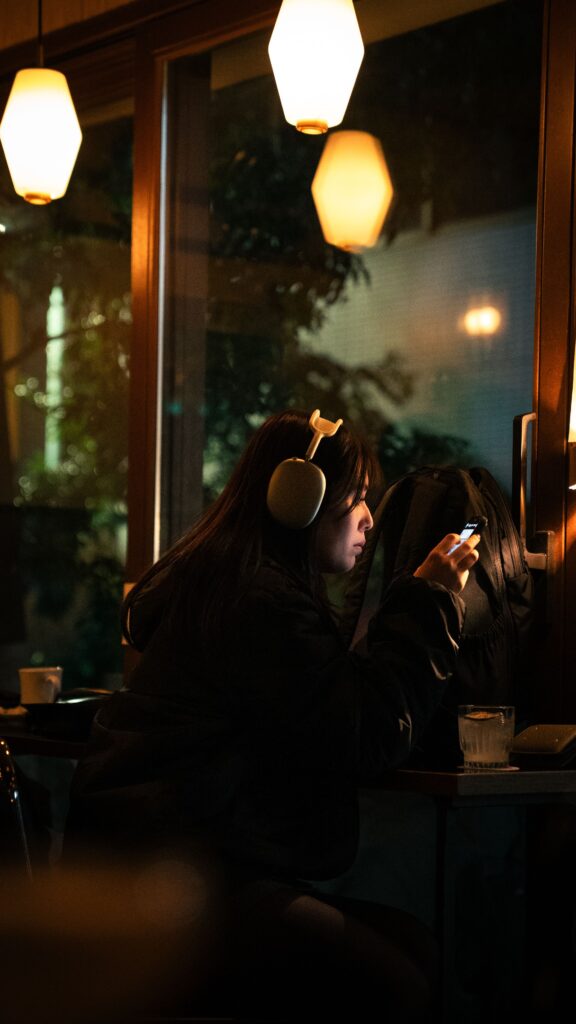 A woman with headphones sitting in a Tokyo cafe, focused on her smartphone in a cozy nighttime setting.