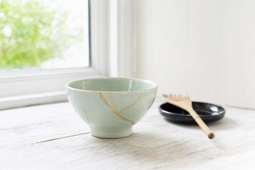 A pale celadon Japanese bowl repaired with gold kintsugi seams on a wooden table, with a lacquer dish and brush nearby
