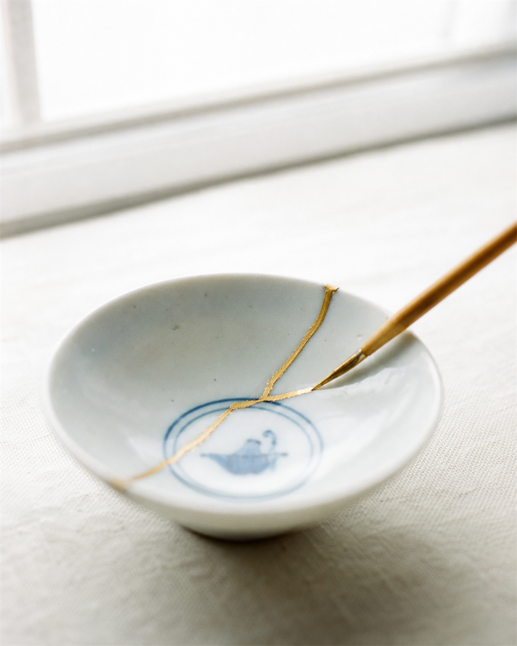 A fine brush laying a gold line along the mended crack of a small white Japanese bowl